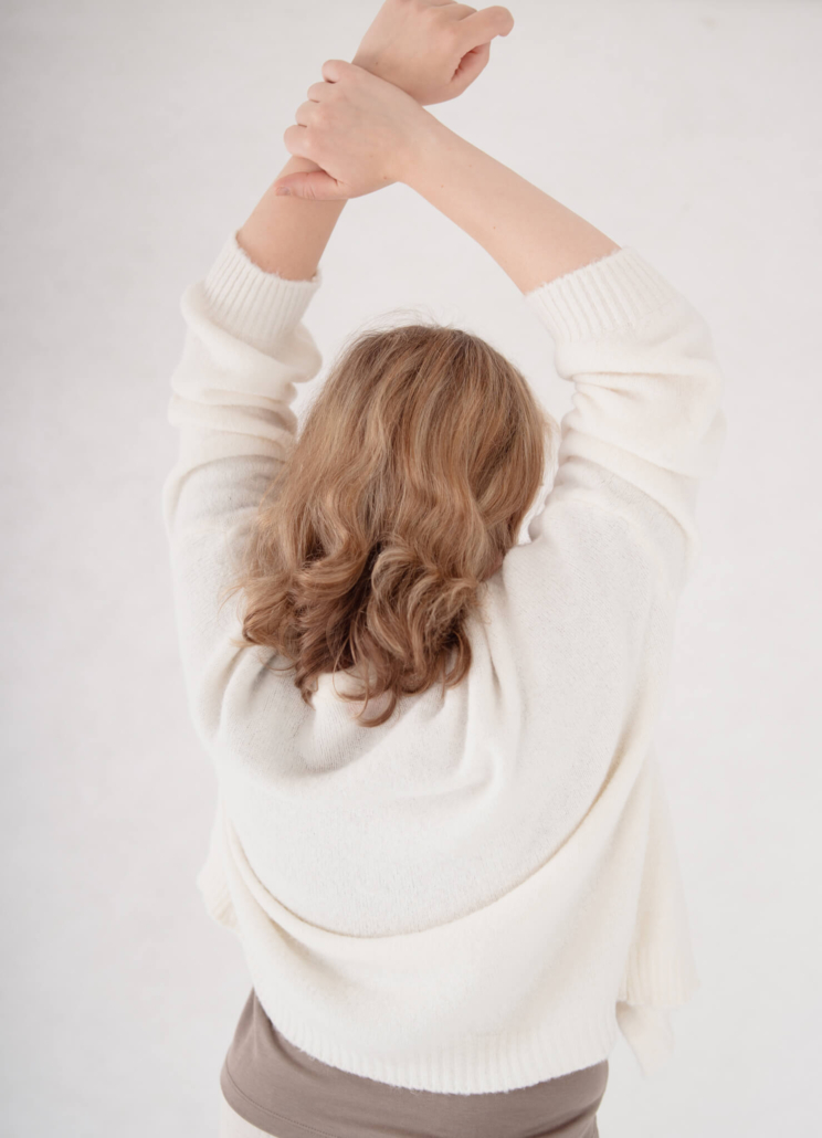 Frau in cremefarbenem Pullover verschränkt entspannt die Arme über dem Kopf – Symbolbild dafür, sich wieder wohl im eigenen Körper zu fühlen.
